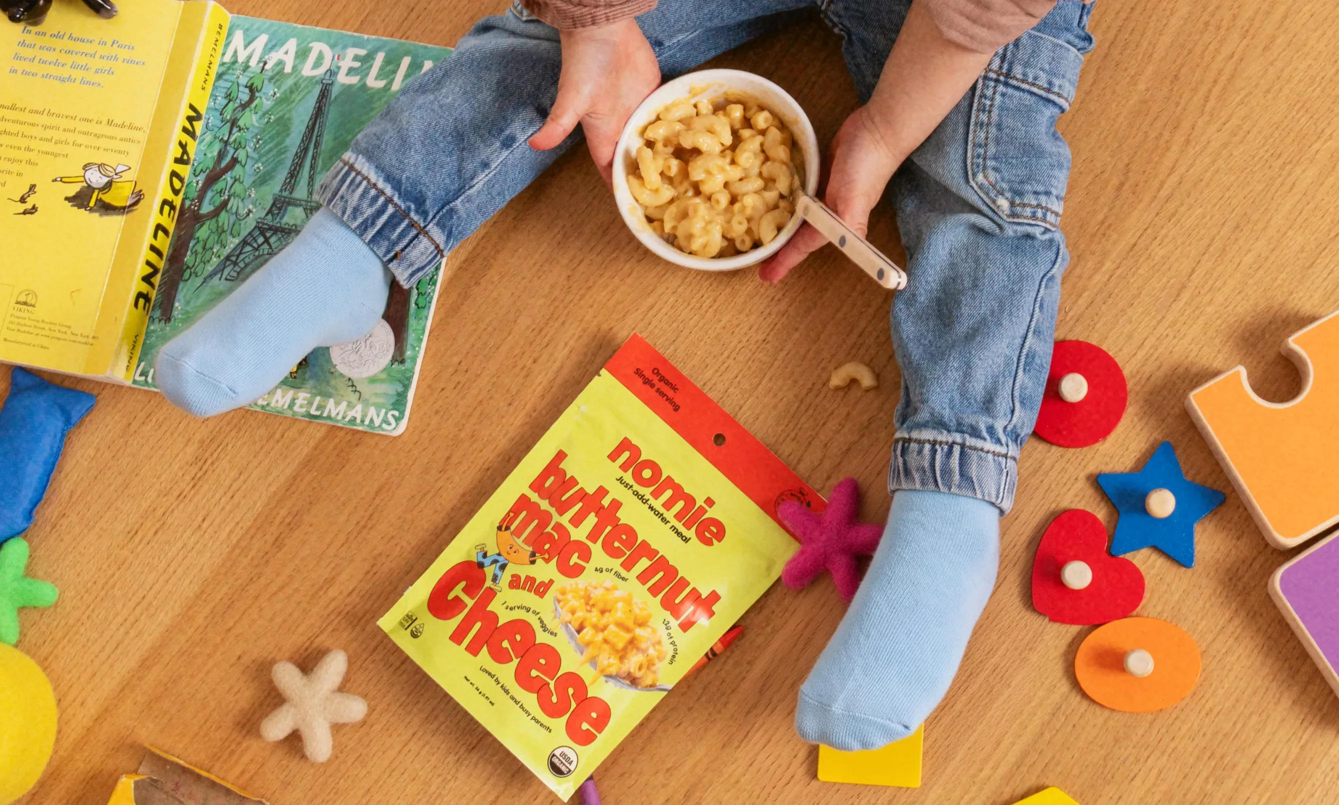 Child on a wooden floor eating Nomie butternut mac and cheese among toys